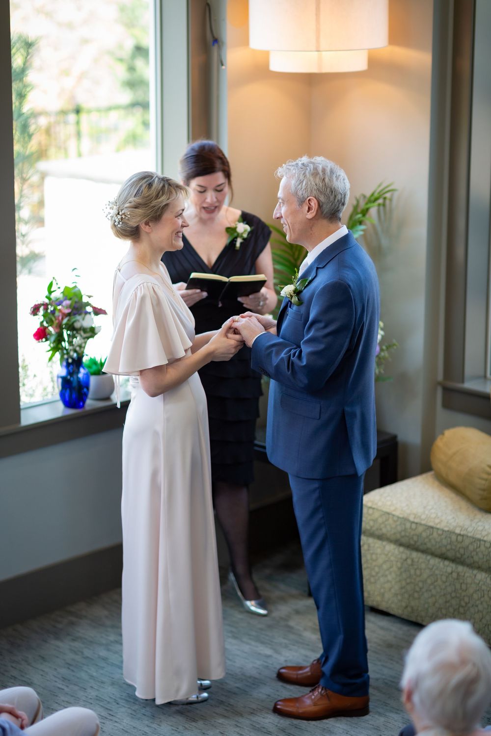 Mature couple exchanging wedding rings during intimate indoor ceremony with officiant holding book.