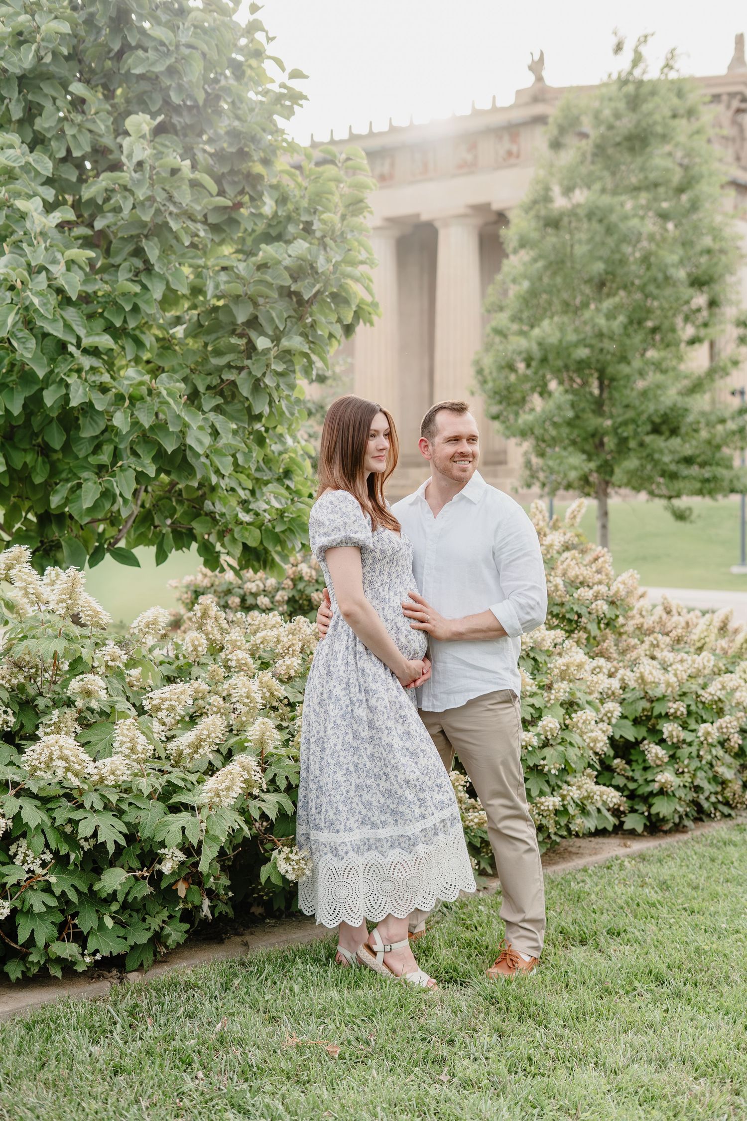 Couple holding hands walking among large columns at a monument, woman in white lace dress touching baby bump.