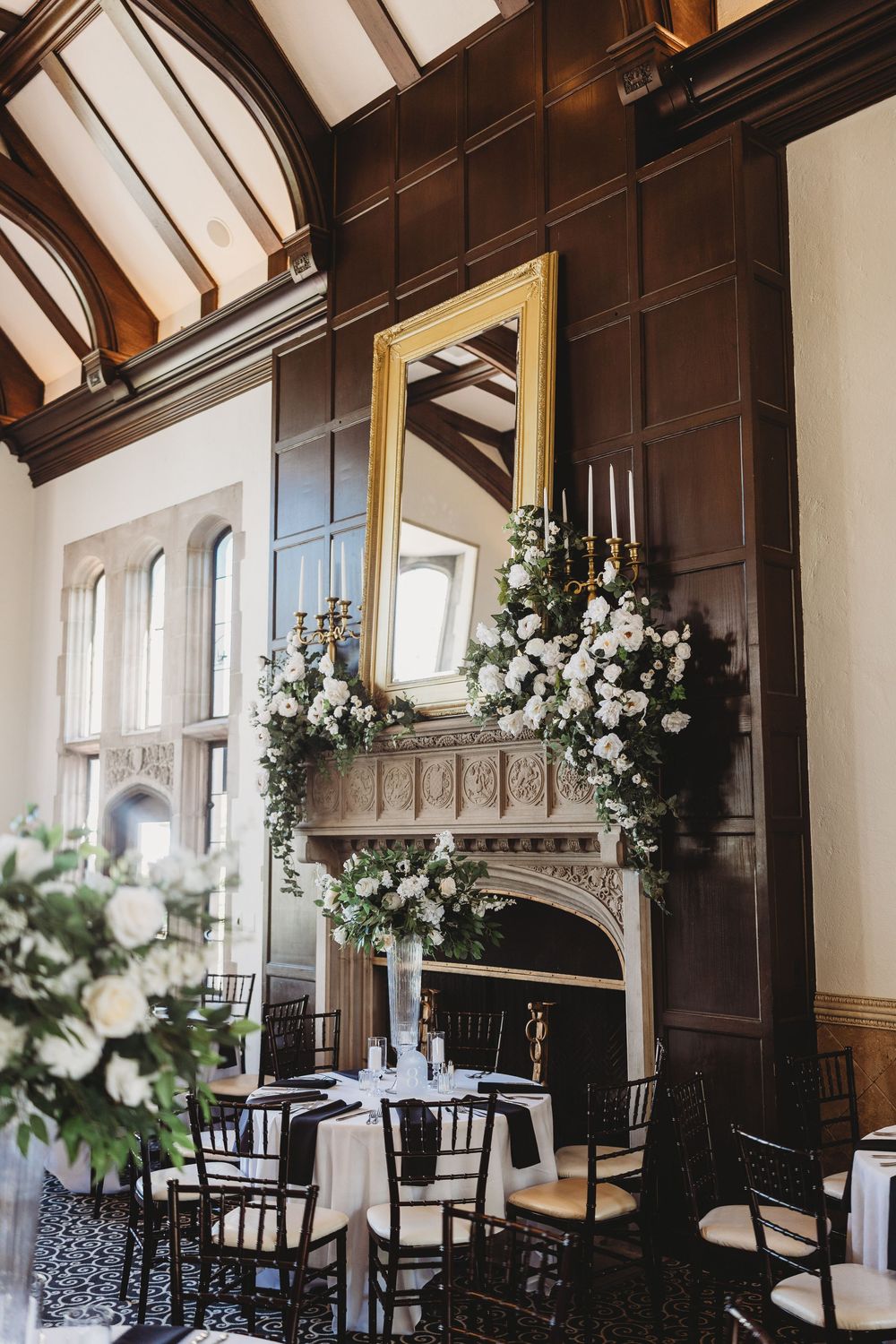 Elegant wedding reception hall with ornate fireplace adorned with white floral arrangements and gold mirror above.