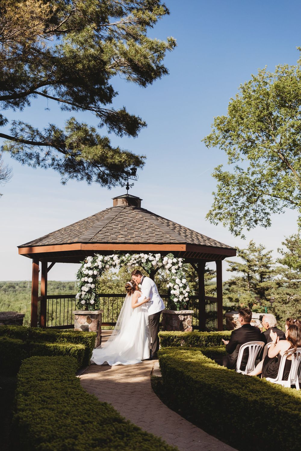 A bride and groom share their first kiss under a wooden gazebo adorned with white flowers at an outdoor ceremony.