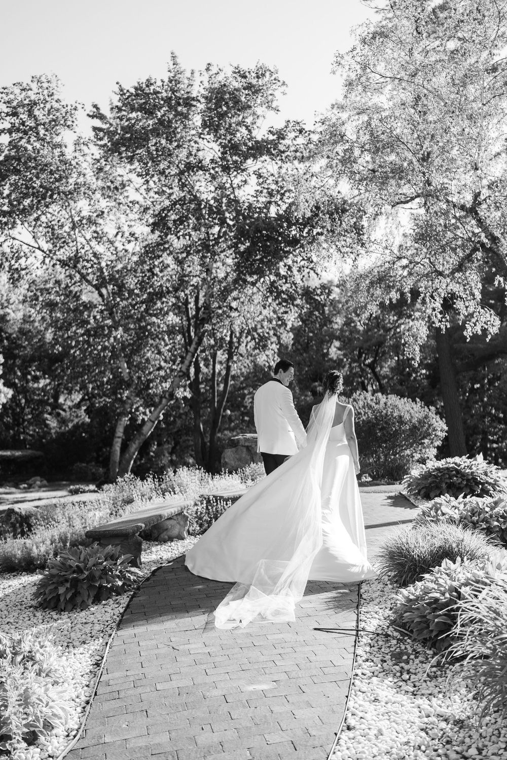 Black and white photo of bride and groom kissing on garden pathway surrounded by lush trees.