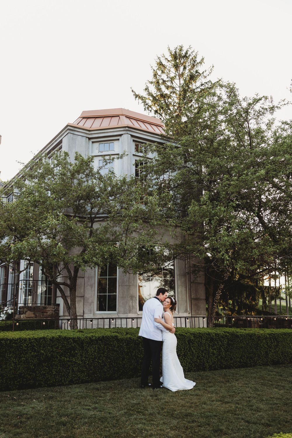 Bride and groom embrace tenderly in front of a grand historic building surrounded by manicured hedges and trees.