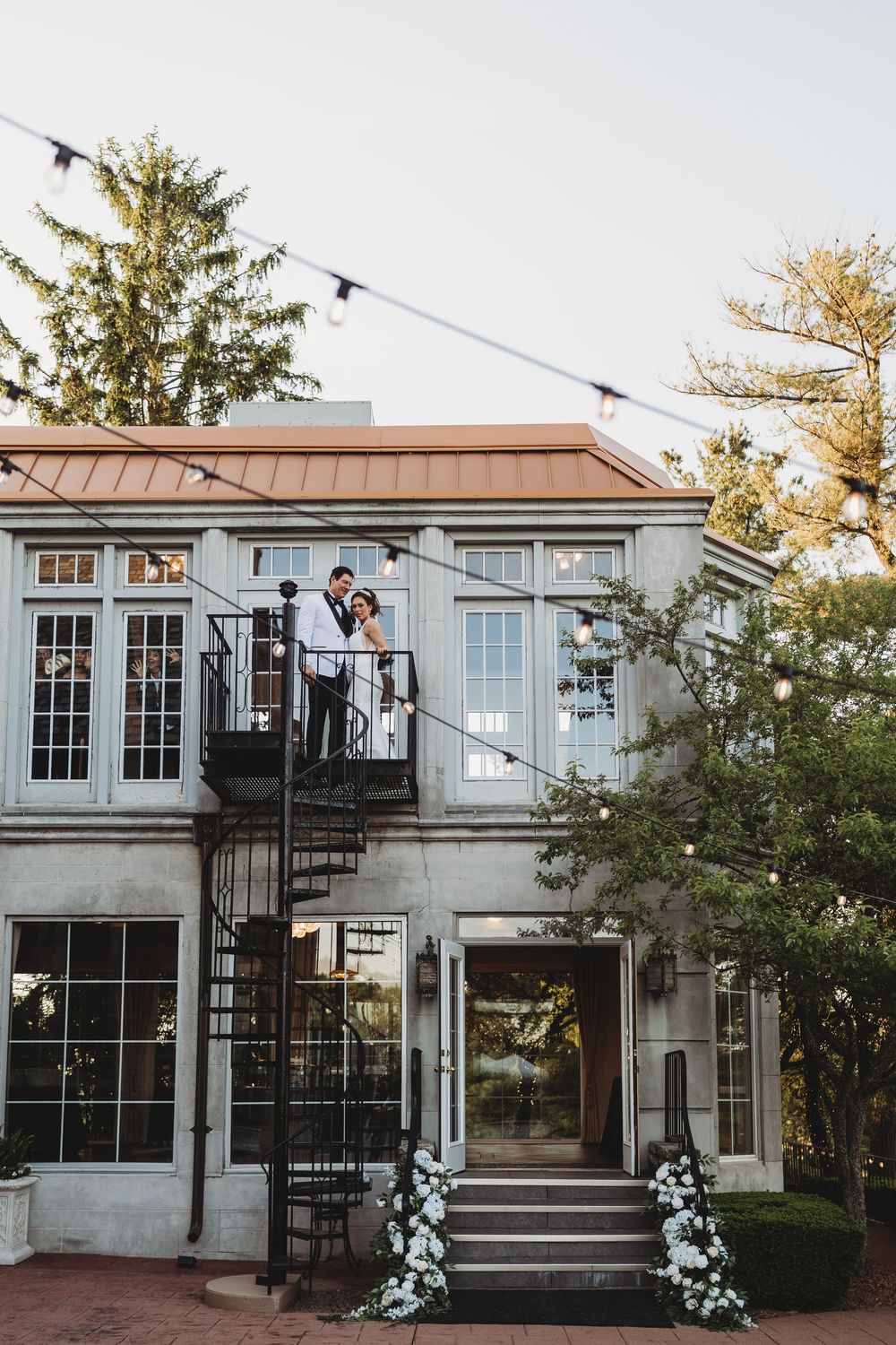 Bride and groom stand on spiral staircase balcony of elegant venue adorned with string lights and white florals.