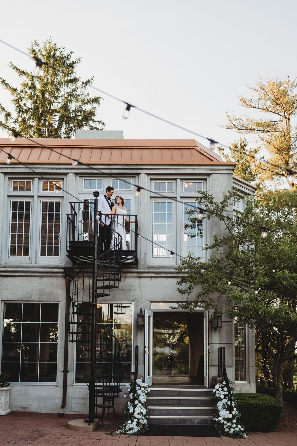 Couple poses on balcony of charming wedding venue with spiral staircase, string lights, and white floral arrangements.