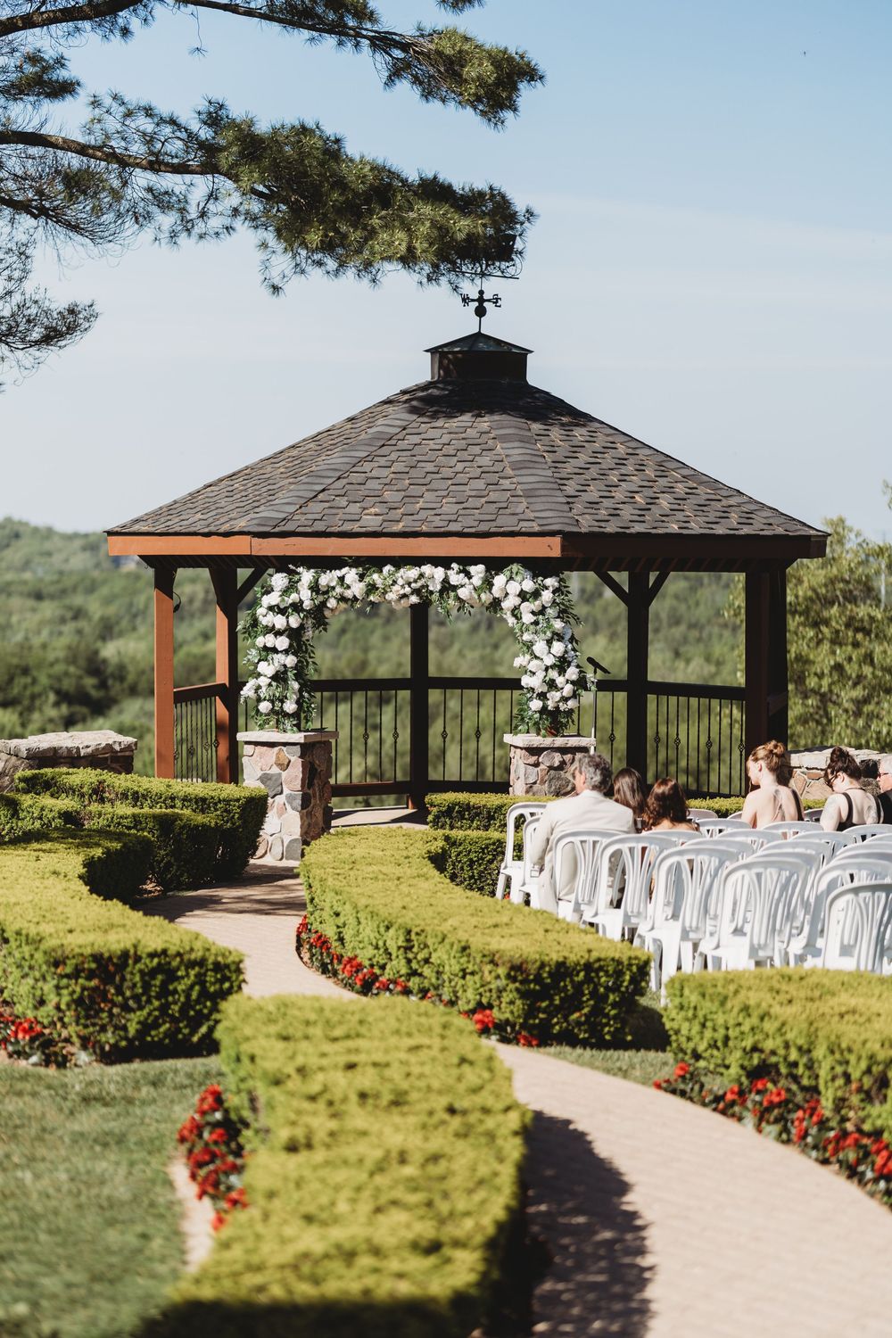 Outdoor wedding ceremony setup at a gazebo decorated with white florals surrounded by manicured hedges and gardens.