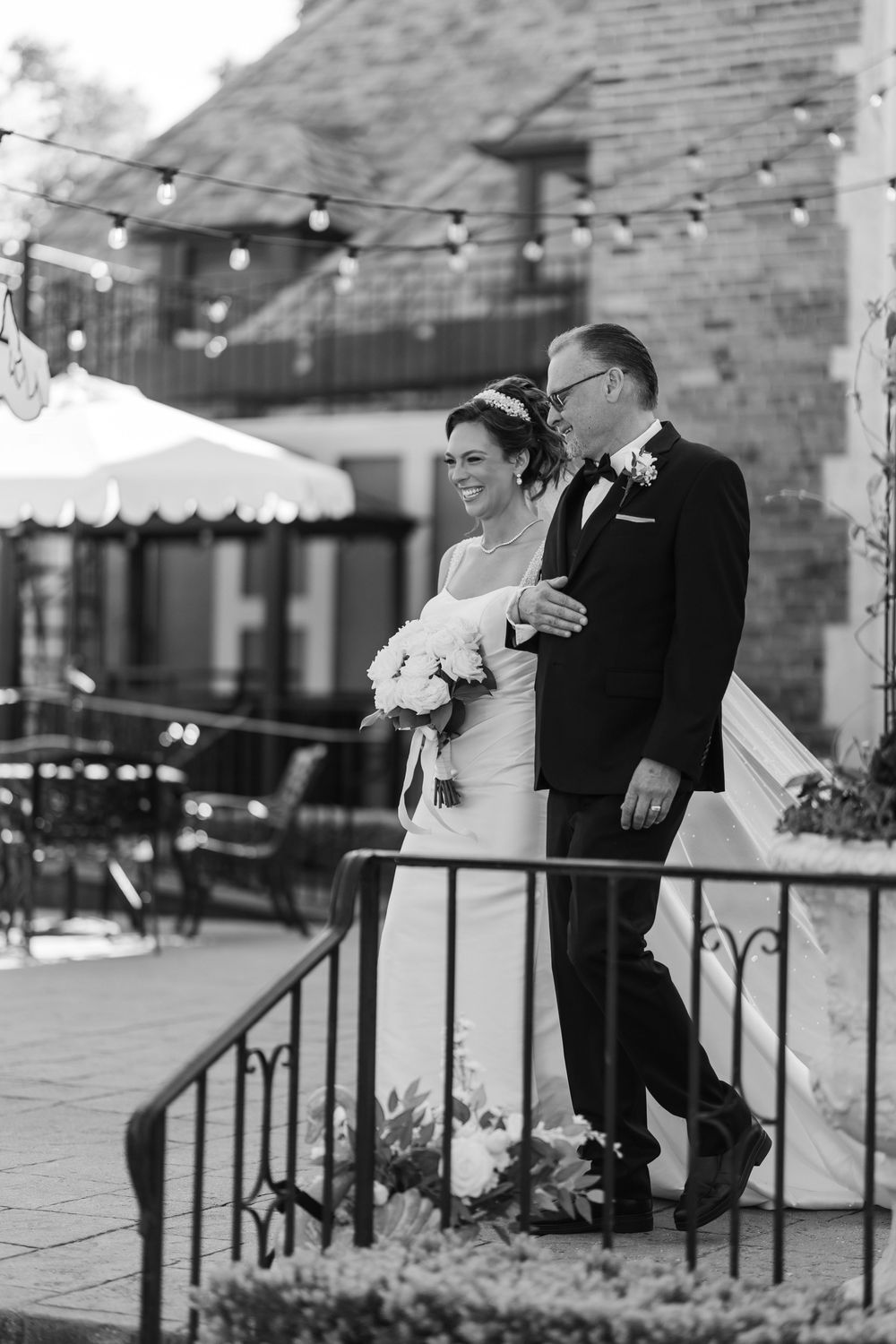 Black and white photo of bride walking with father down staircase under string lights holding white bouquet.