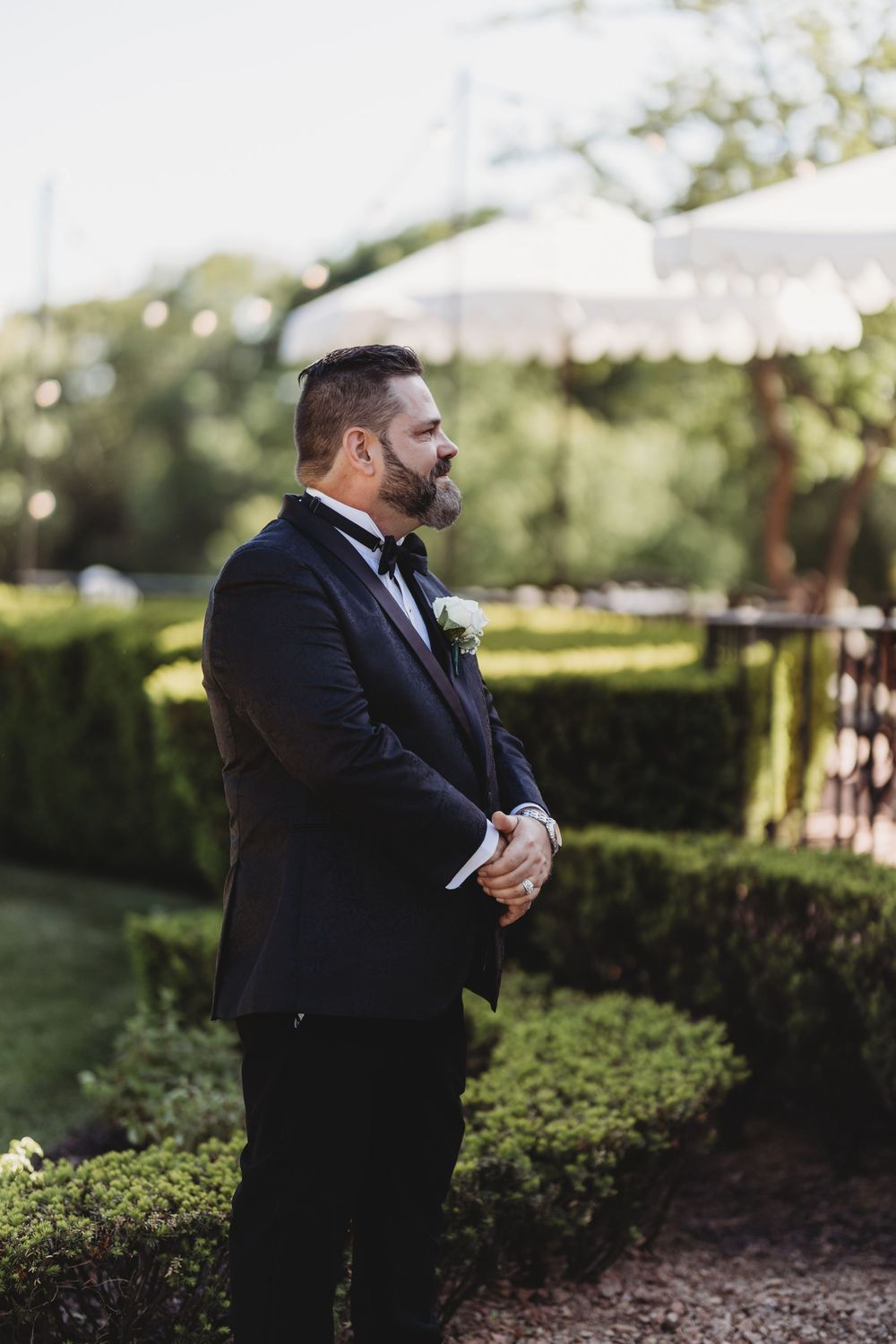 Dapper groom in navy tuxedo with boutonniere standing in manicured garden before wedding ceremony.