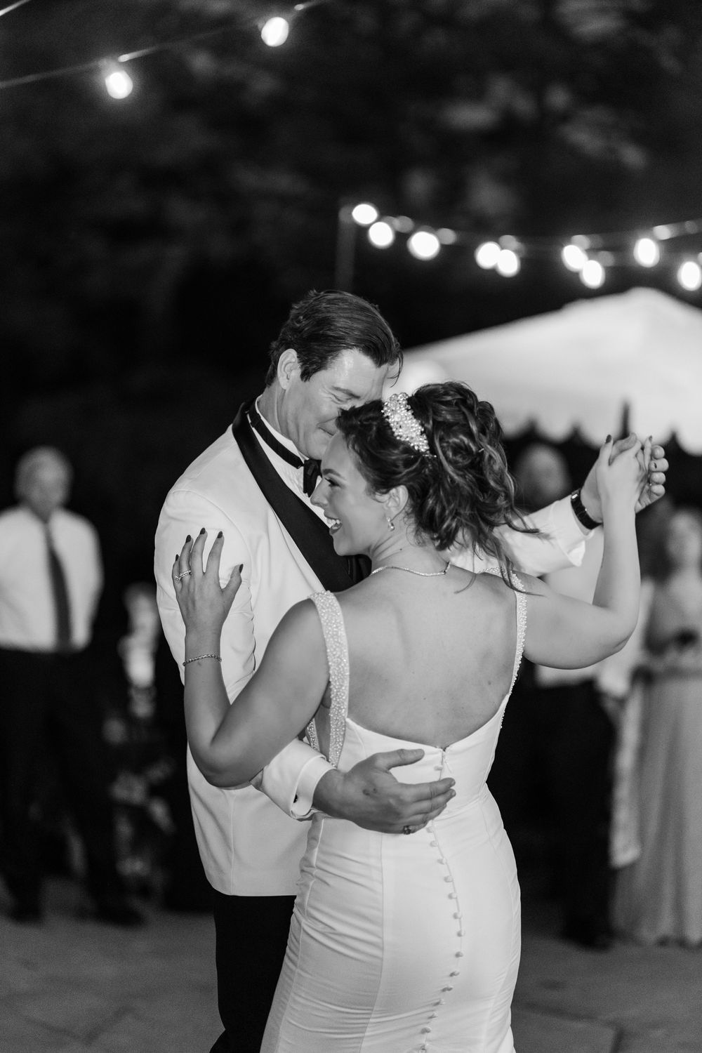 Black and white photo of bride and groom sharing their first dance under string lights at an outdoor wedding reception.
