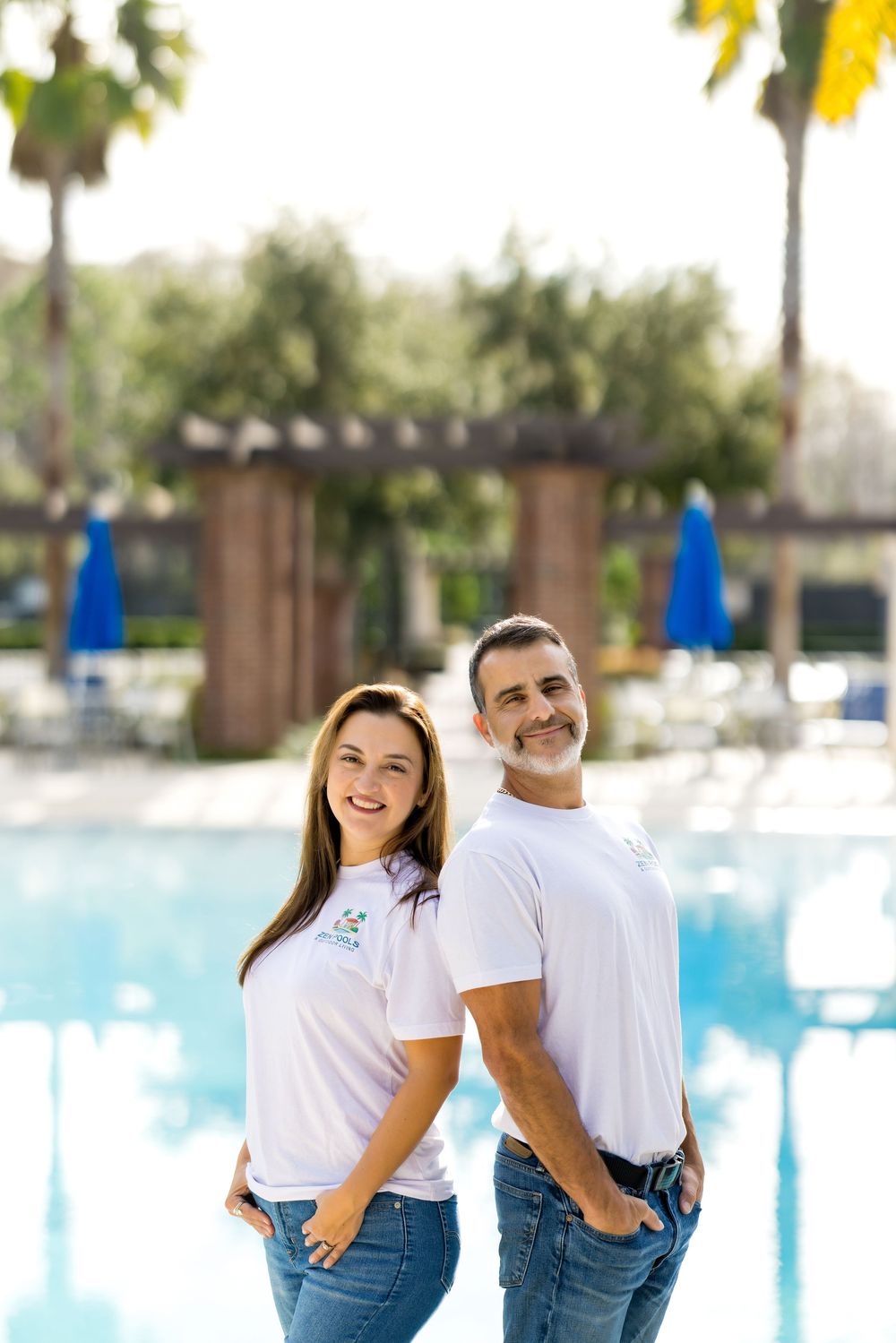 A smiling couple in matching white t-shirts and jeans pose back-to-back near a resort swimming pool.