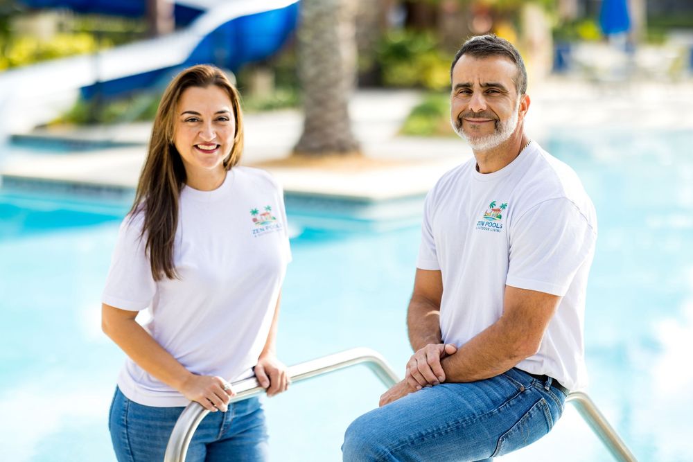 Two smiling pool service professionals in white branded shirts pose by a sunny outdoor swimming pool.