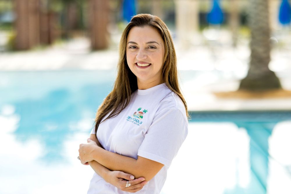 Young woman in white polo shirt smiling with arms crossed beside a sparkling outdoor swimming pool.