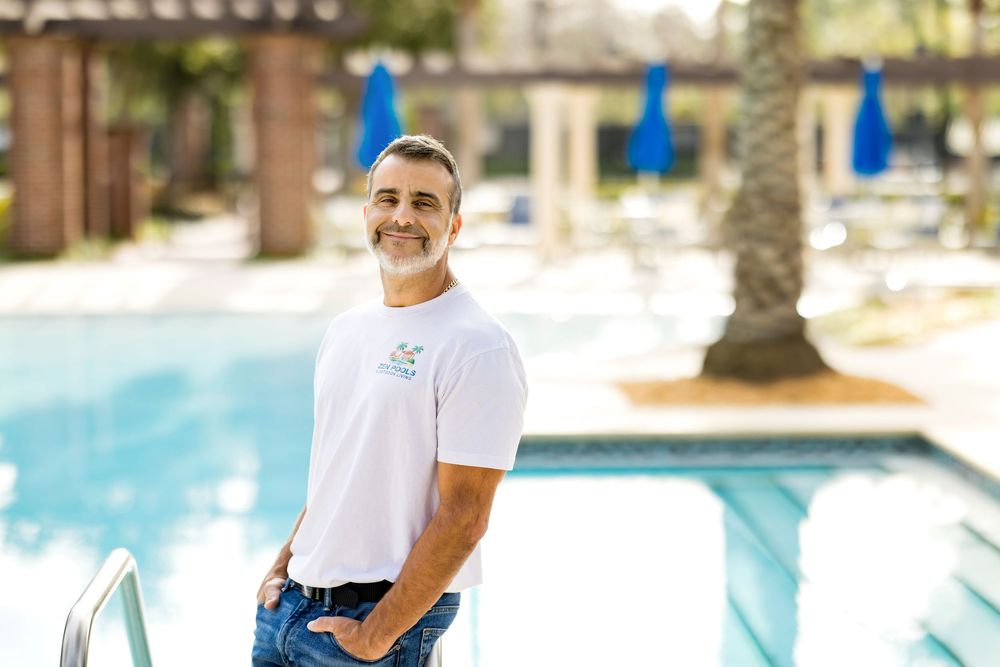 Man in white pool service shirt smiling poolside with blue umbrellas and palm trees in background.