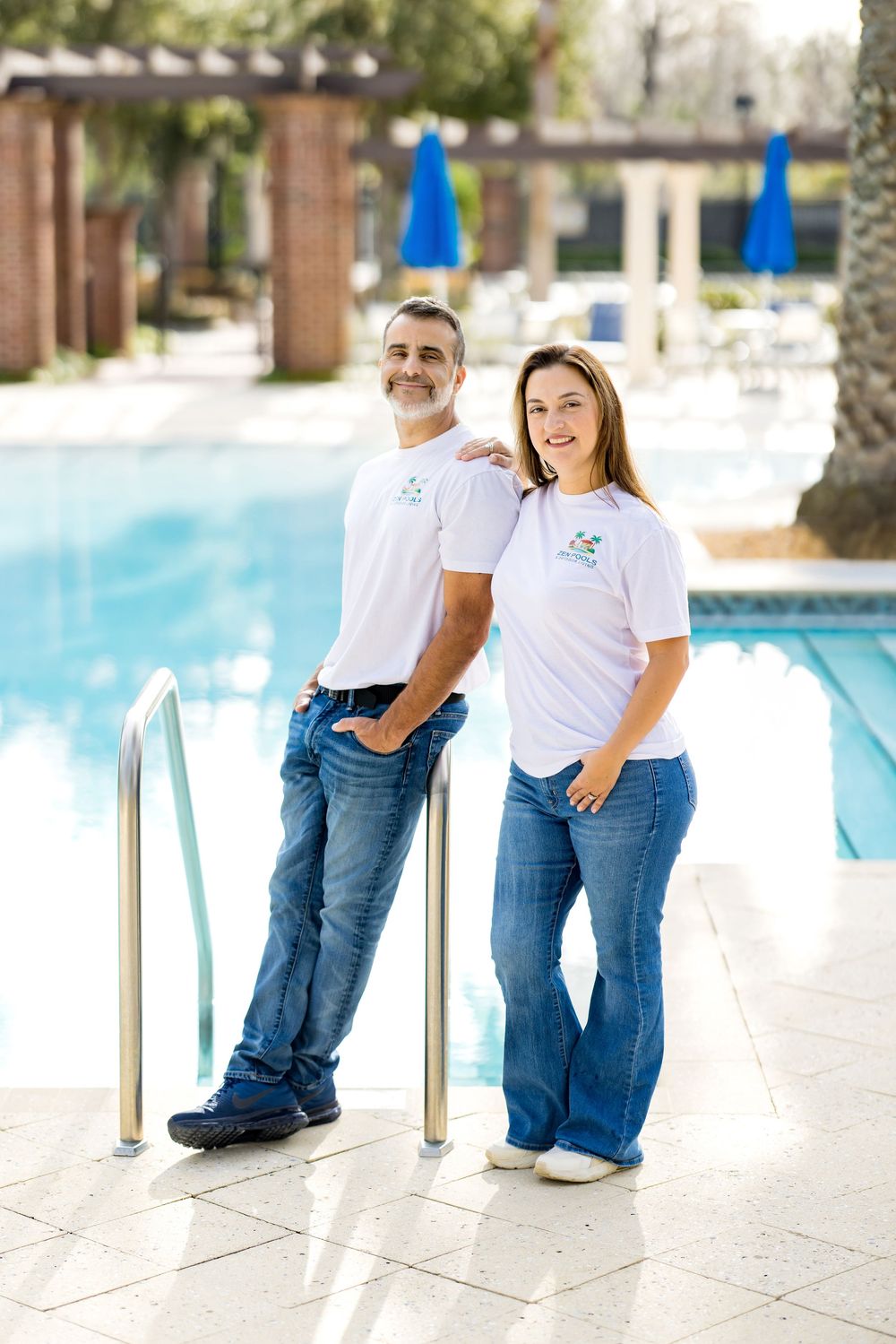 A man and woman in matching white shirts and jeans pose smiling by a sparkling outdoor swimming pool.
