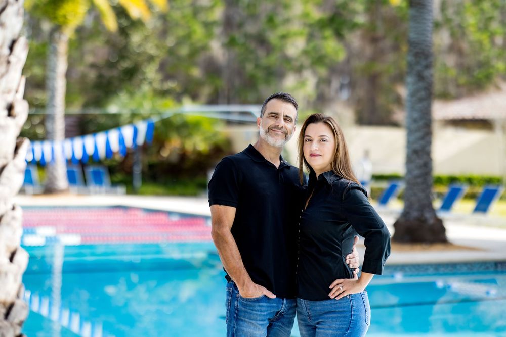 Couple in matching black shirts posing together in front of a sunny outdoor swimming pool.