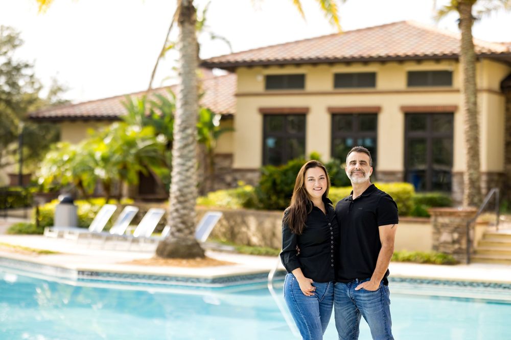 Couple in matching black outfits posing together in front of a resort pool with palm trees.