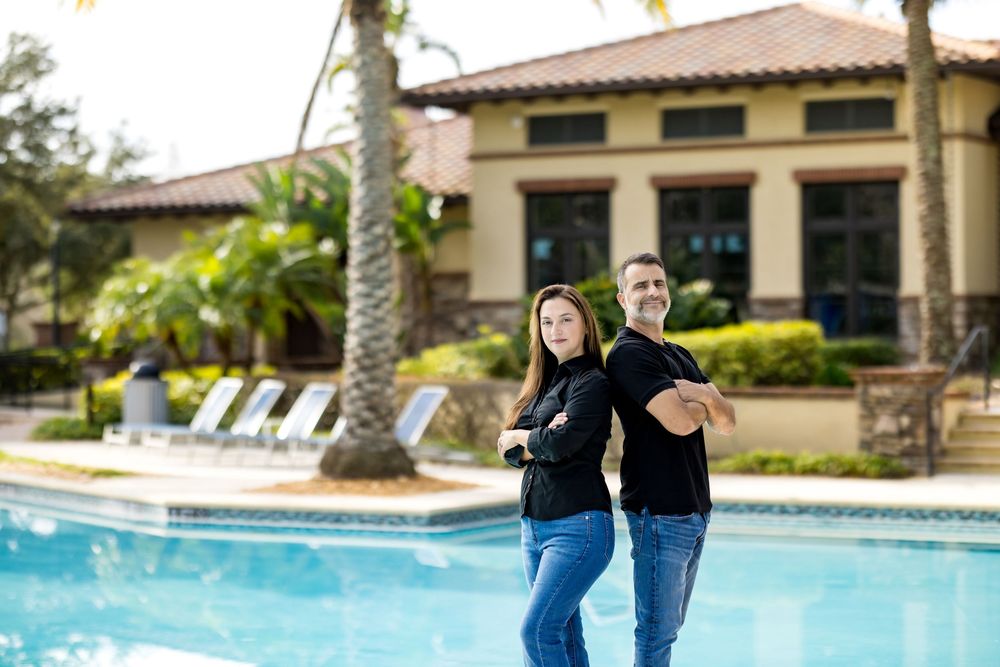 Couple standing back-to-back smiling by a resort swimming pool surrounded by palm trees.