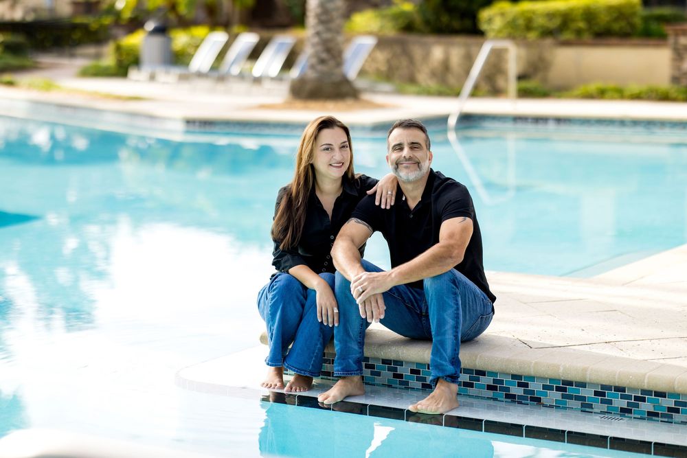 A smiling couple sits together by a resort swimming pool, dressed casually in black tops and jeans on a sunny day.