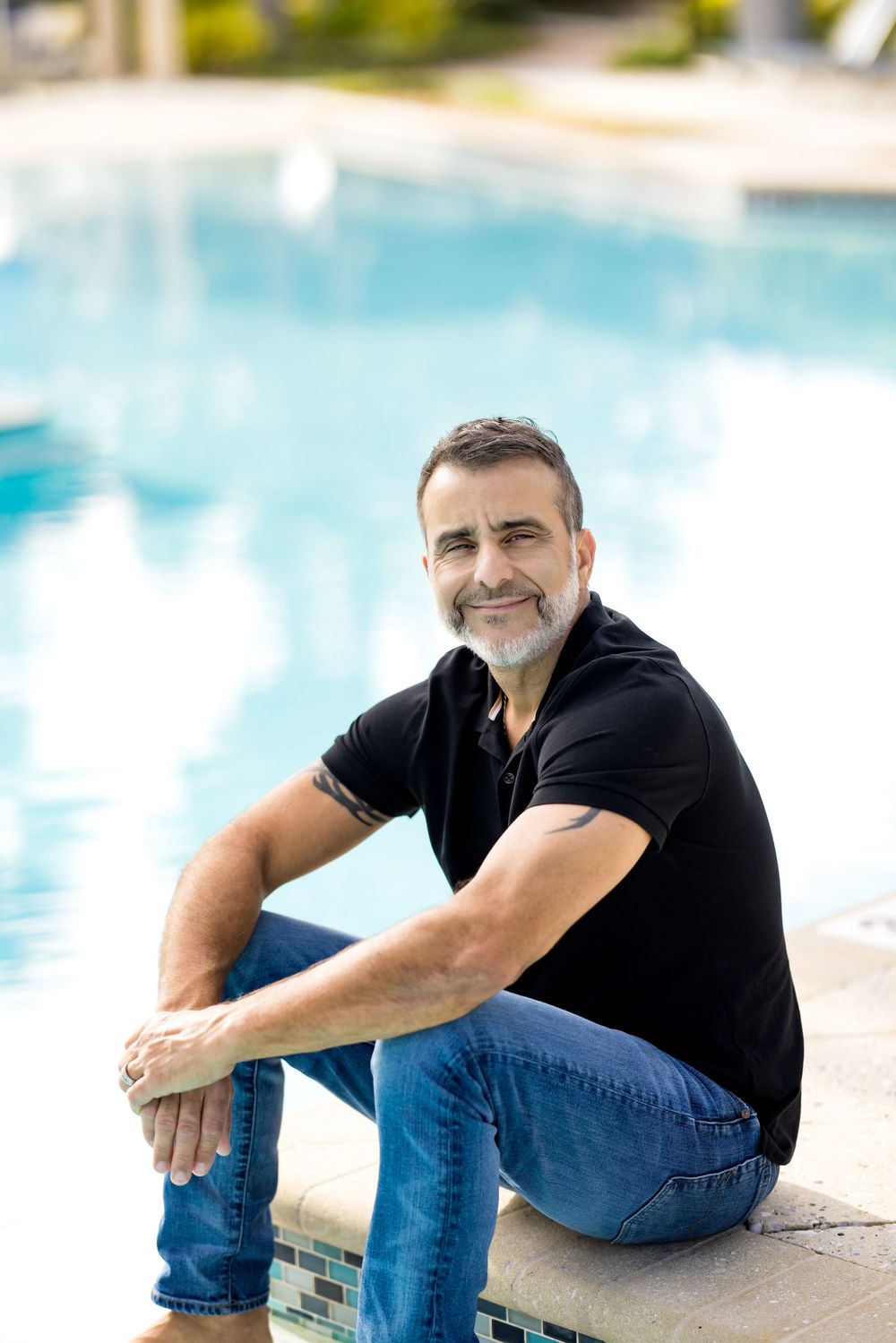 Middle-aged man with gray beard sitting poolside in black polo shirt and jeans, smiling outdoors.