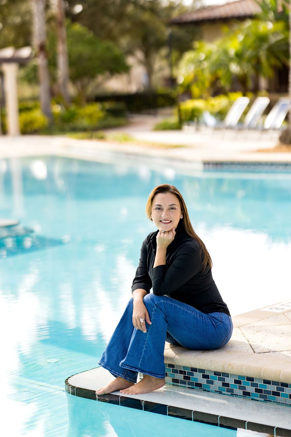 Woman in black top and jeans sitting poolside at a resort, smiling at camera on a sunny day.
