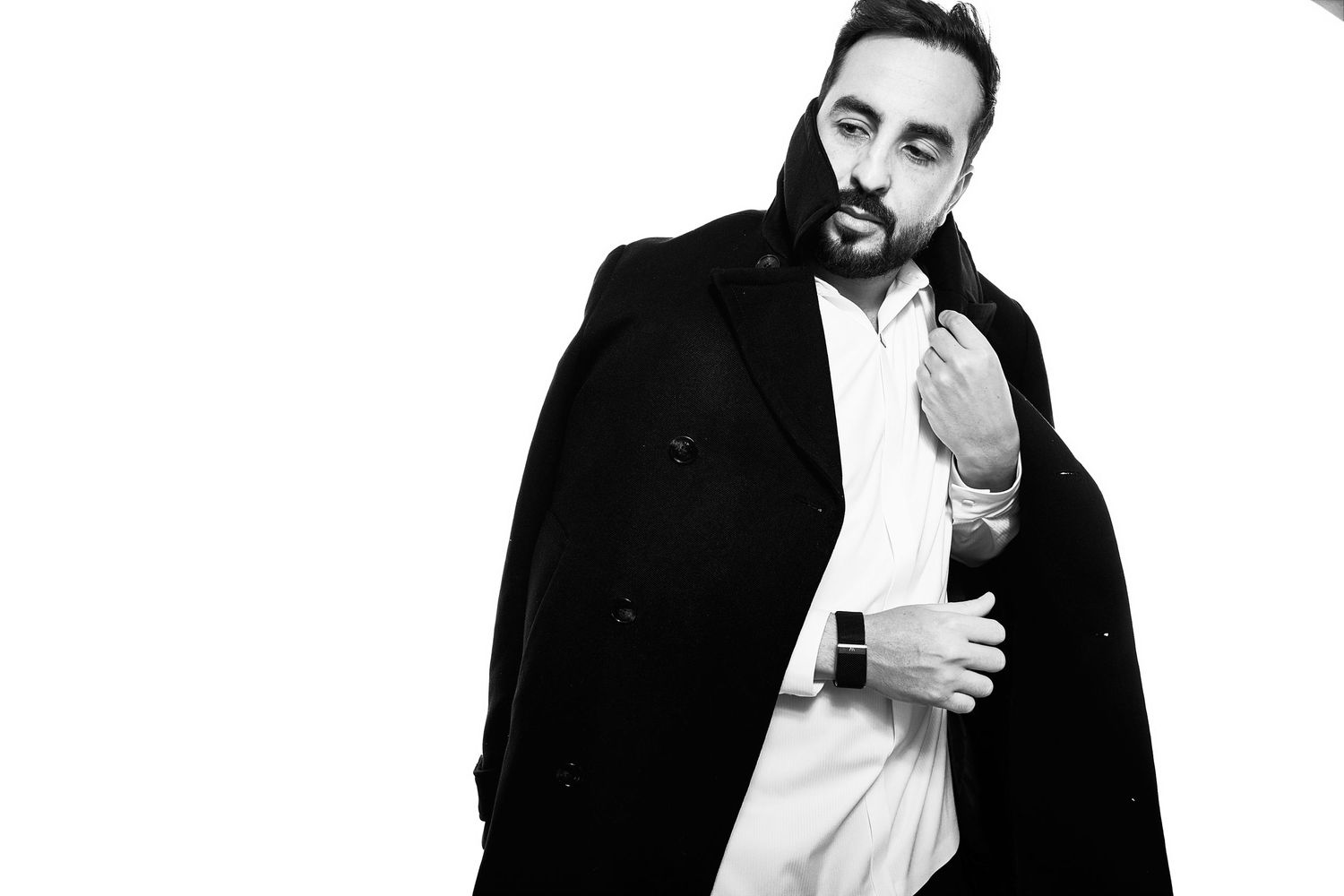 Black and white portrait of a bearded man in a white shirt and black coat posing confidently against white background.