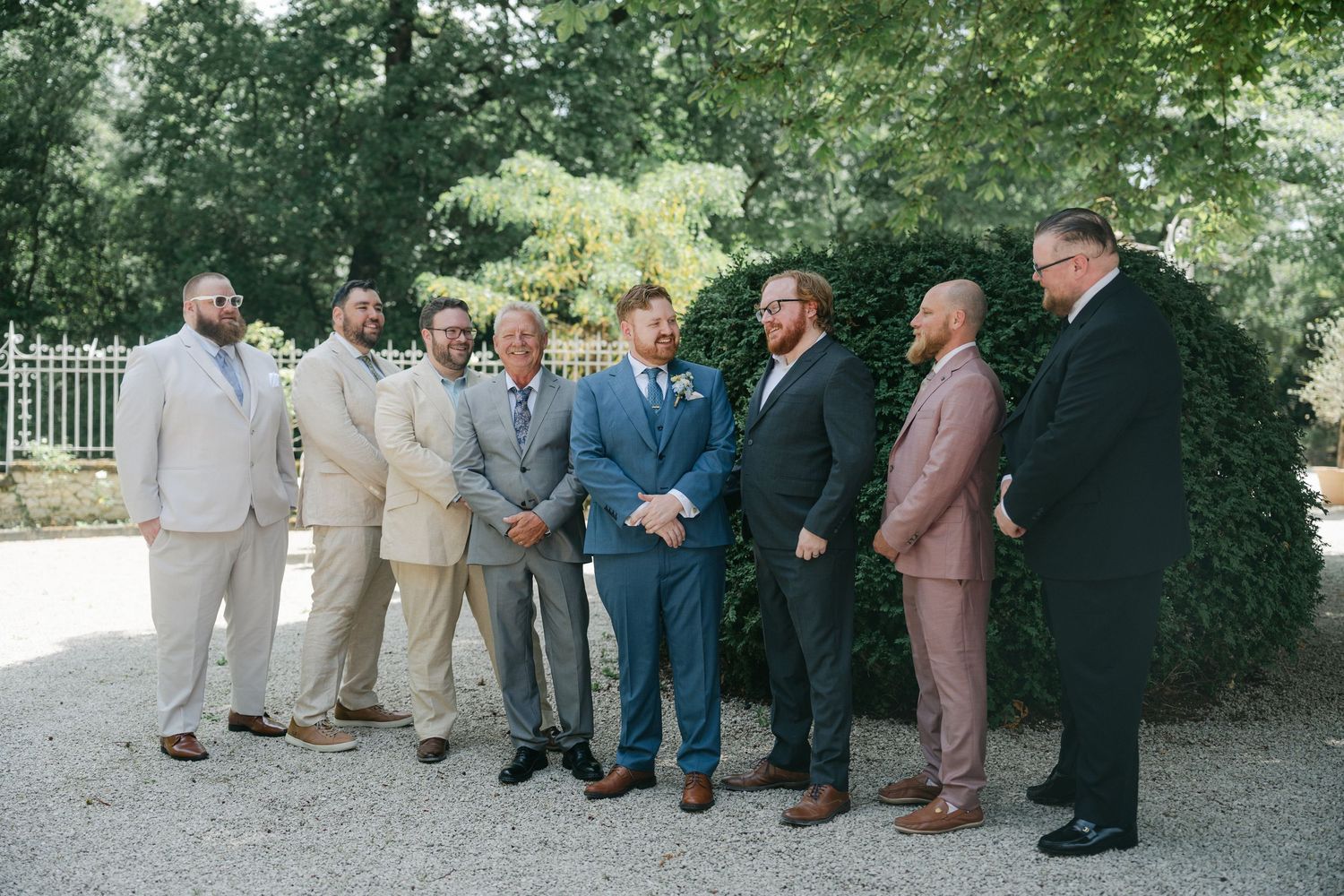 A groom and his groomsmen pose outdoors in various suits before a wedding ceremony surrounded by lush green trees.
