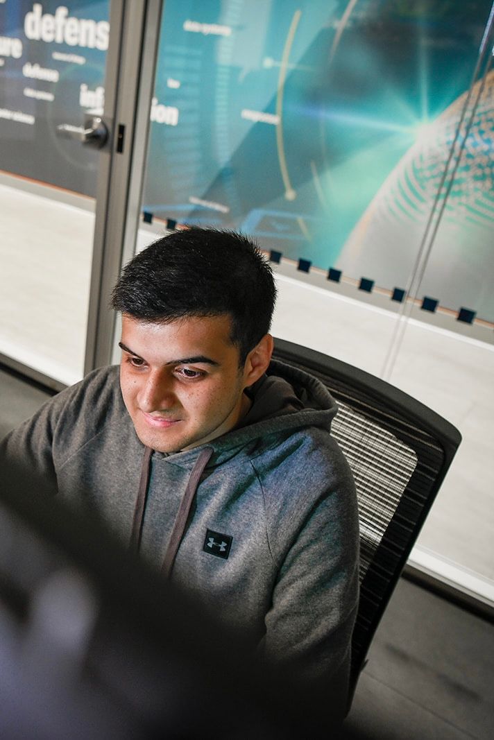 Young man in gray hoodie working at computer in modern office with cybersecurity themed display wall.