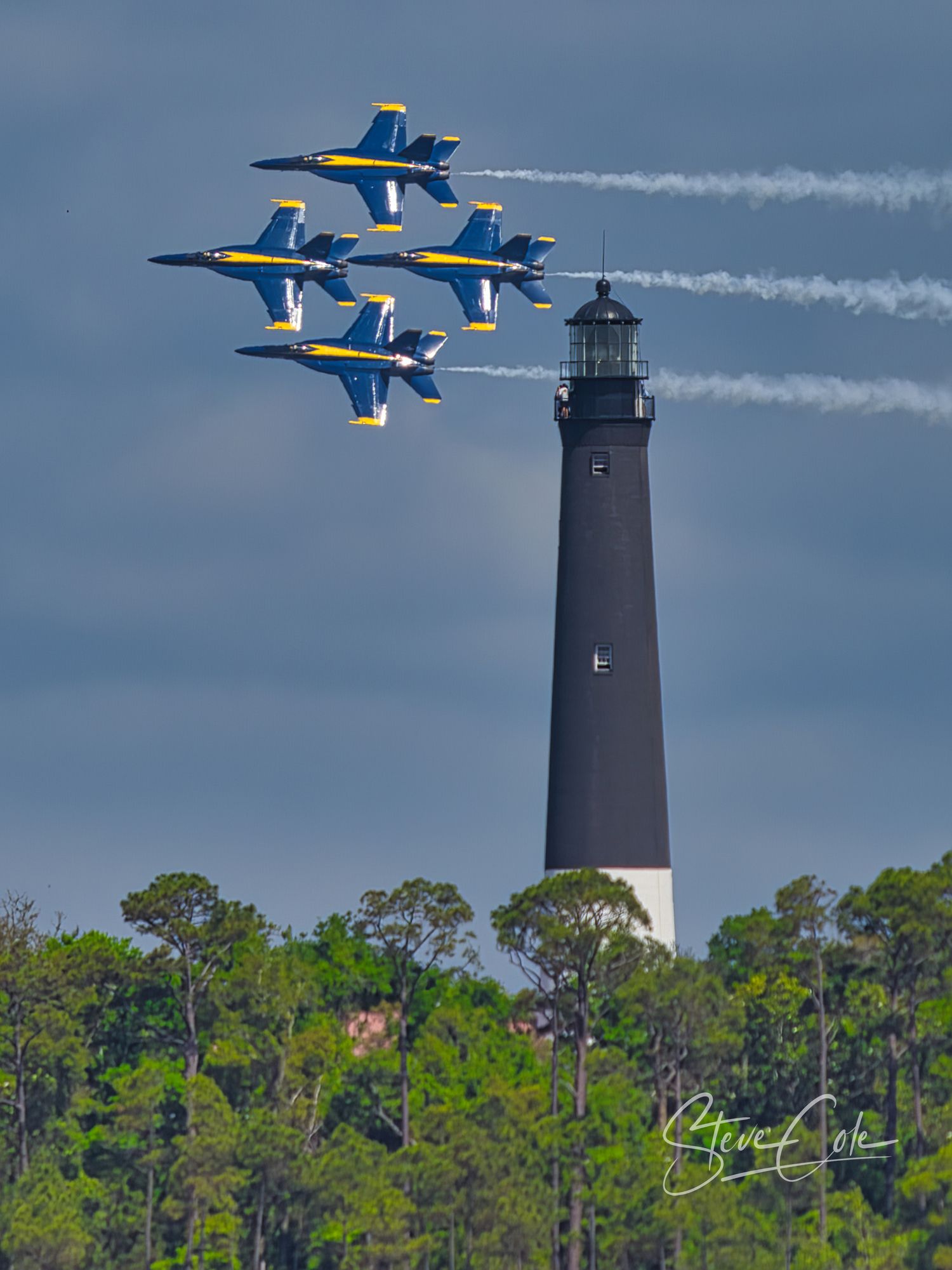 Four Blue Angels jets in diamond formation flying past a historic coastal lighthouse, fine art photography by Steve Cole.
