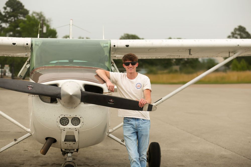Young man in sunglasses leaning on a small Cessna propeller plane on a tarmac airfield.