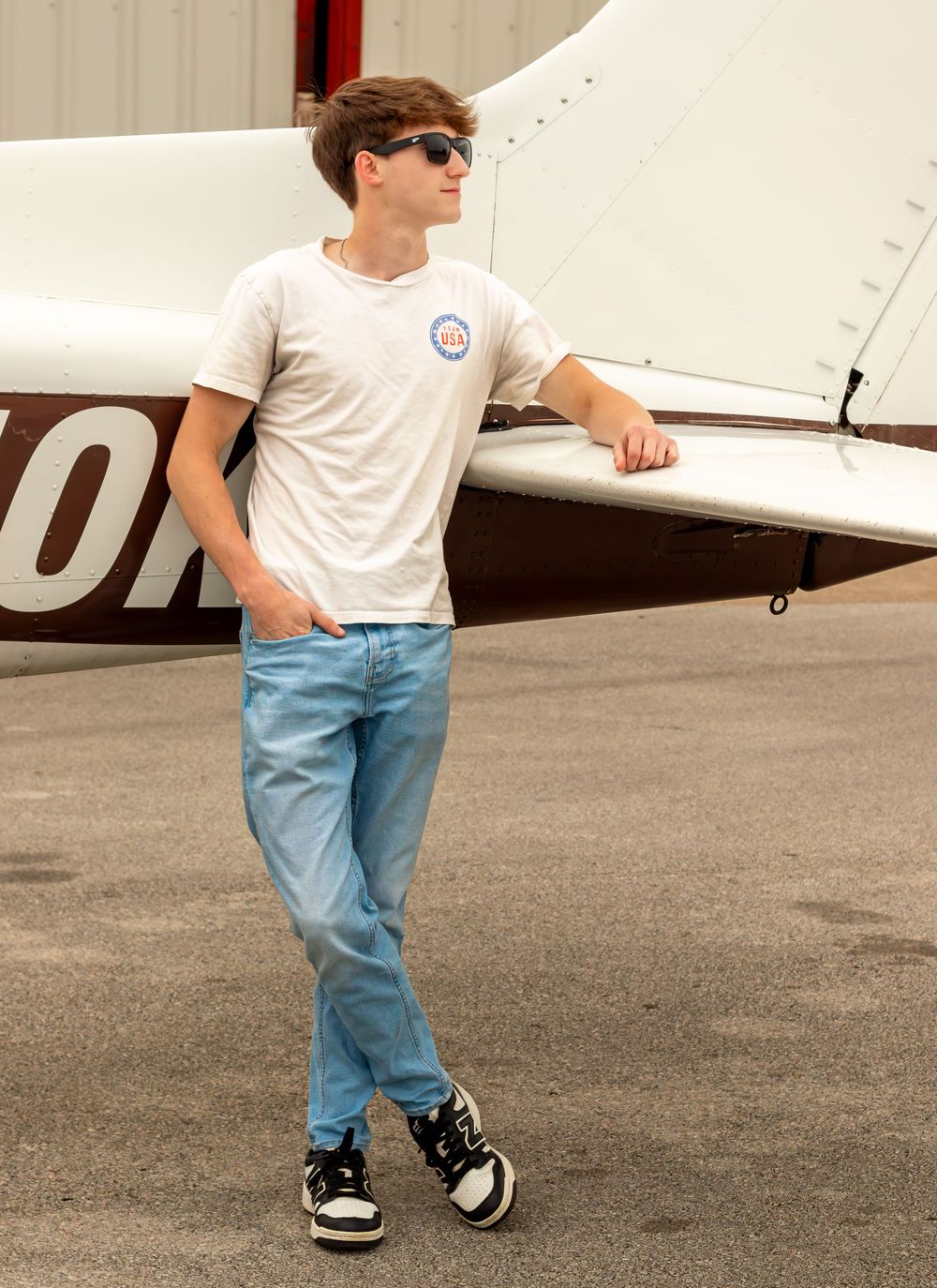 Teen boy in white t-shirt, jeans, and sunglasses leaning against a small aircraft on a tarmac.