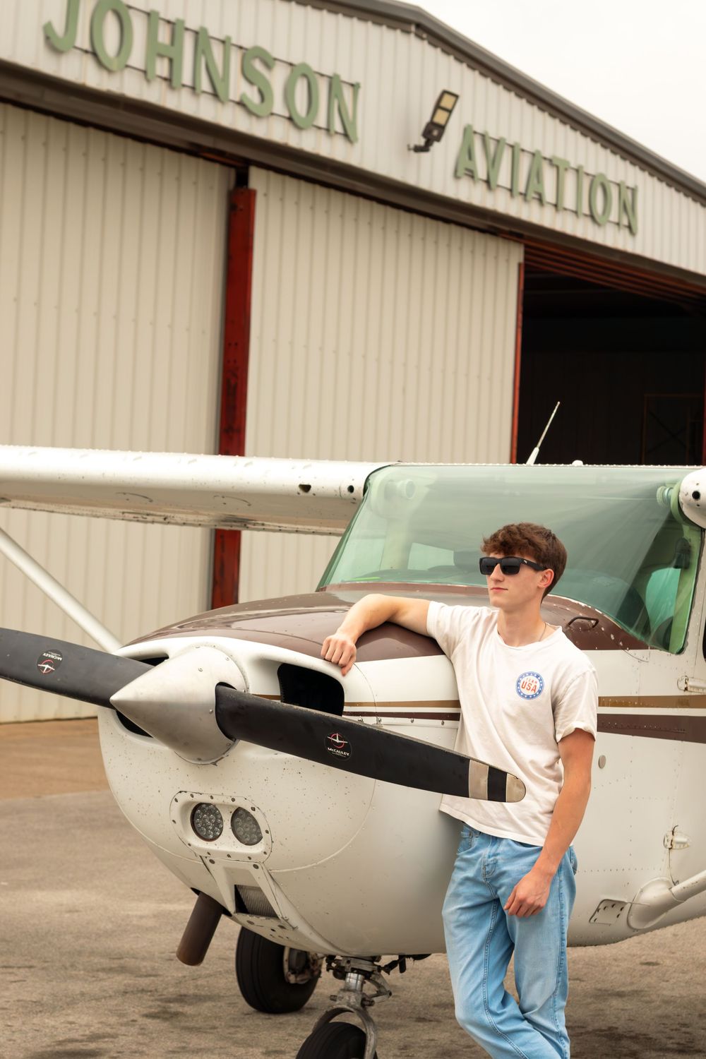 Young man in sunglasses leaning on a small Cessna aircraft outside Johnson Aviation hangar.
