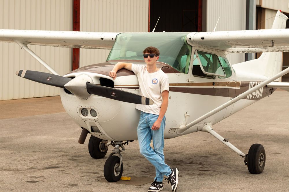 Young man in sunglasses posing with a small Cessna aircraft on an airport tarmac near a hangar.