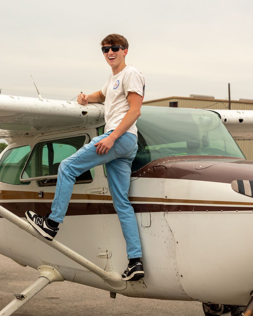 Young man in sunglasses sitting on a small Cessna airplane wing at an airfield, wearing jeans and a white t-shirt.