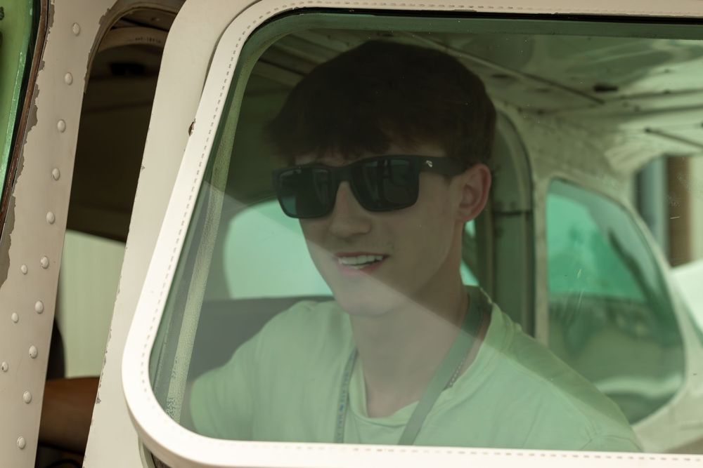Young man wearing sunglasses smiling inside a small aircraft cockpit window.