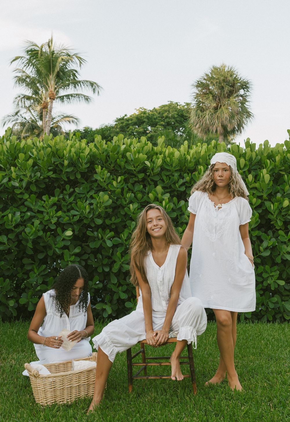 Group wearing white summer dresses relaxing on grass with palm trees and greenery in background.