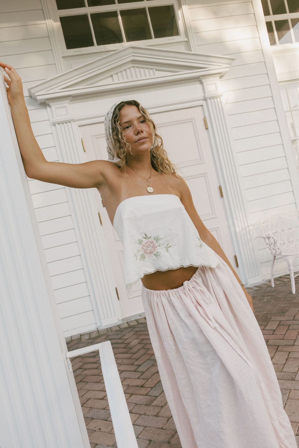 A woman in a flowing white summer dress poses elegantly against a white colonial-style building exterior.