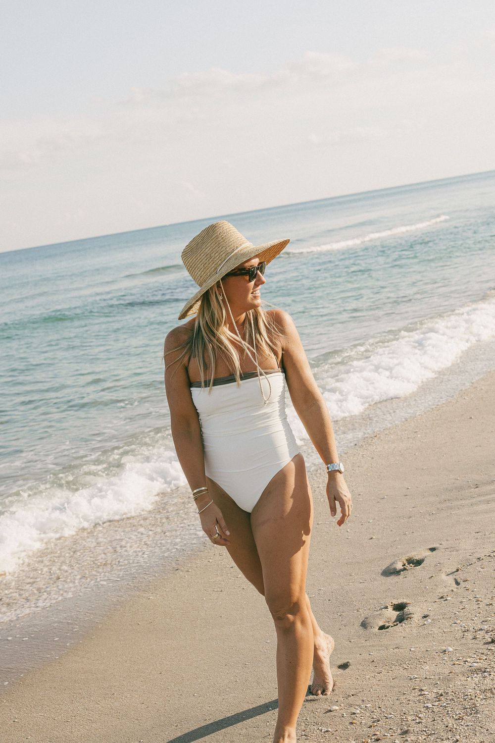 A person in a white swimsuit and straw hat strolls along a sunny beach collecting seashells by the ocean.