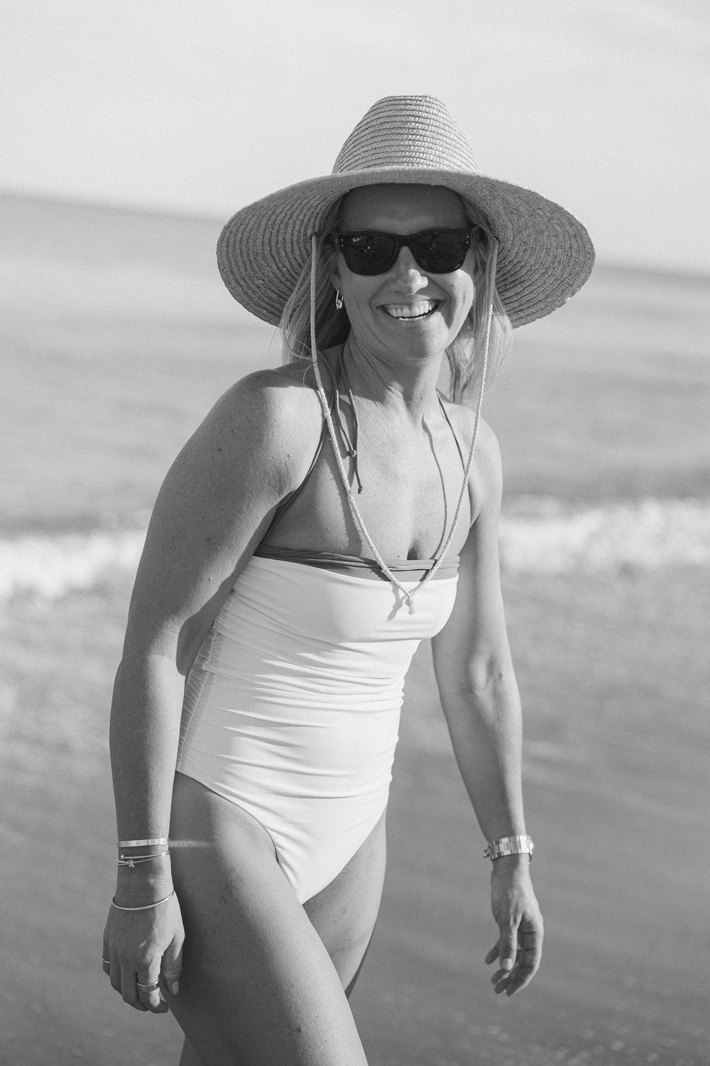 A person in a white swimsuit and sun hat enjoys a sunny day at the beach in black and white photography.