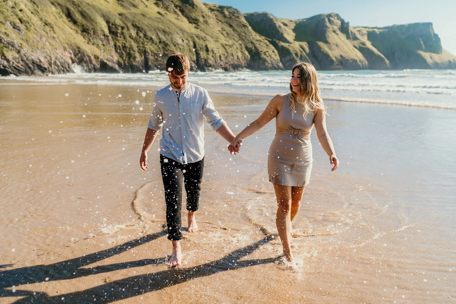 Couple holding hands walking on a sandy beach with ocean waves and green cliffs in the background.