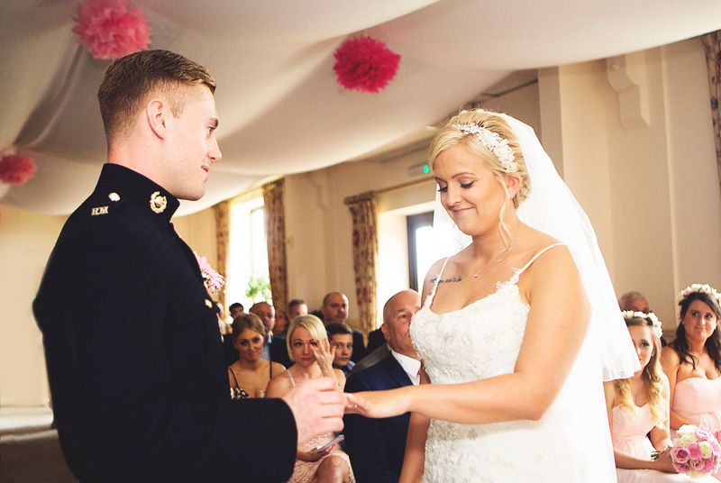 Military groom and bride exchanging rings during wedding ceremony with bridesmaids seated in background. Surrey wedding photos