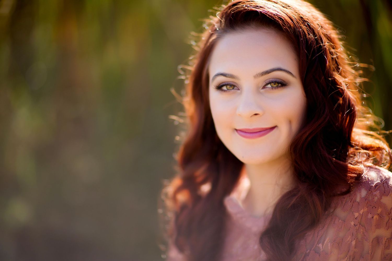 A portrait of a woman with long wavy brown hair wearing a white top against a soft blurred natural background.
