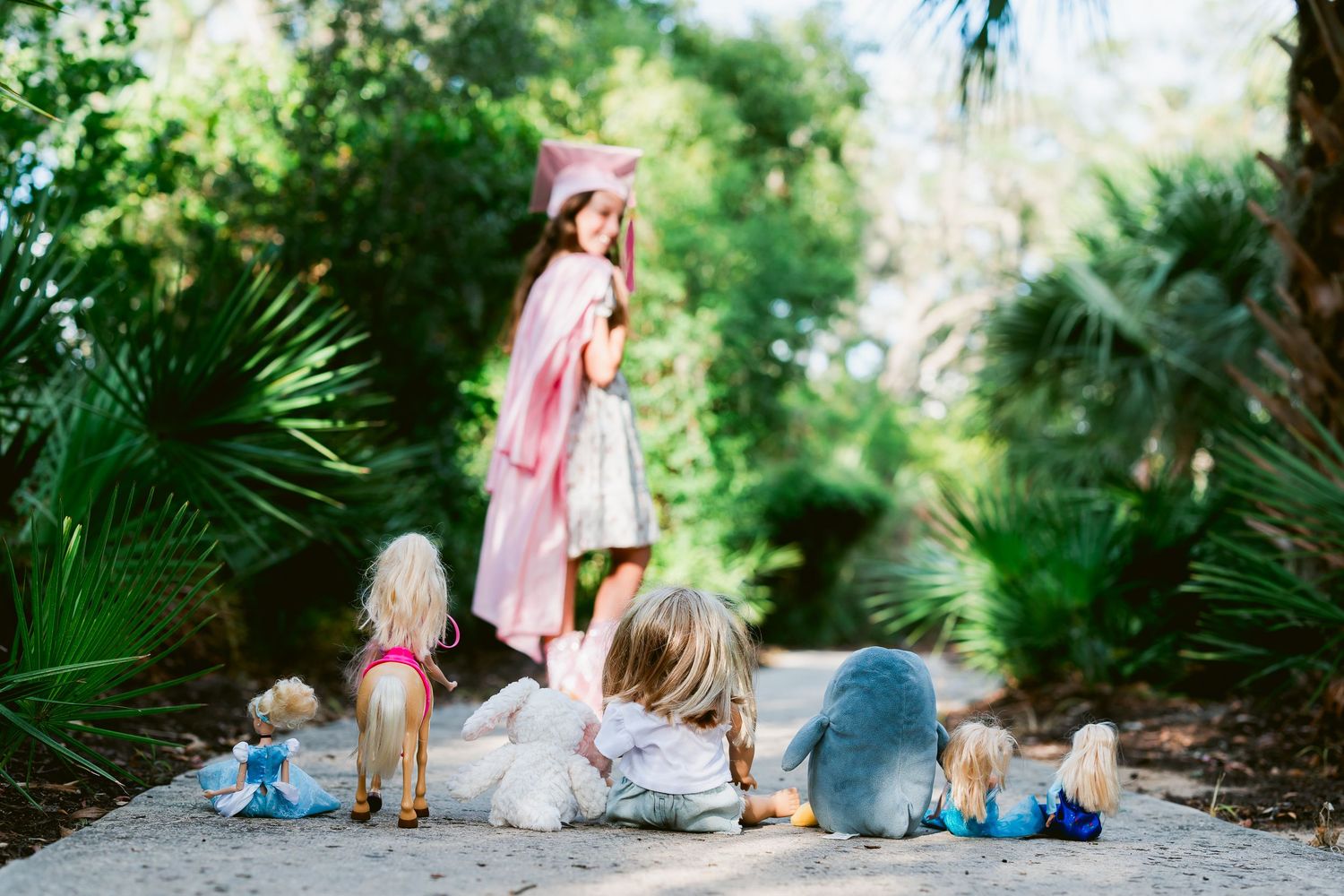 Girl in pink graduation cap and gown stands on garden path with younger children and dolls sitting behind her.