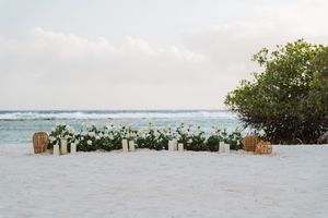 White floral arrangement with greenery on white sand beach at sunset, perfect for a coastal wedding ceremony.