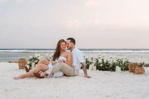 Romantic couple sitting together on white sand beach at sunset with floral decorations nearby.