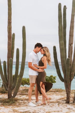 Couple embracing between tall cacti on a sandy beach with turquoise ocean in the background.