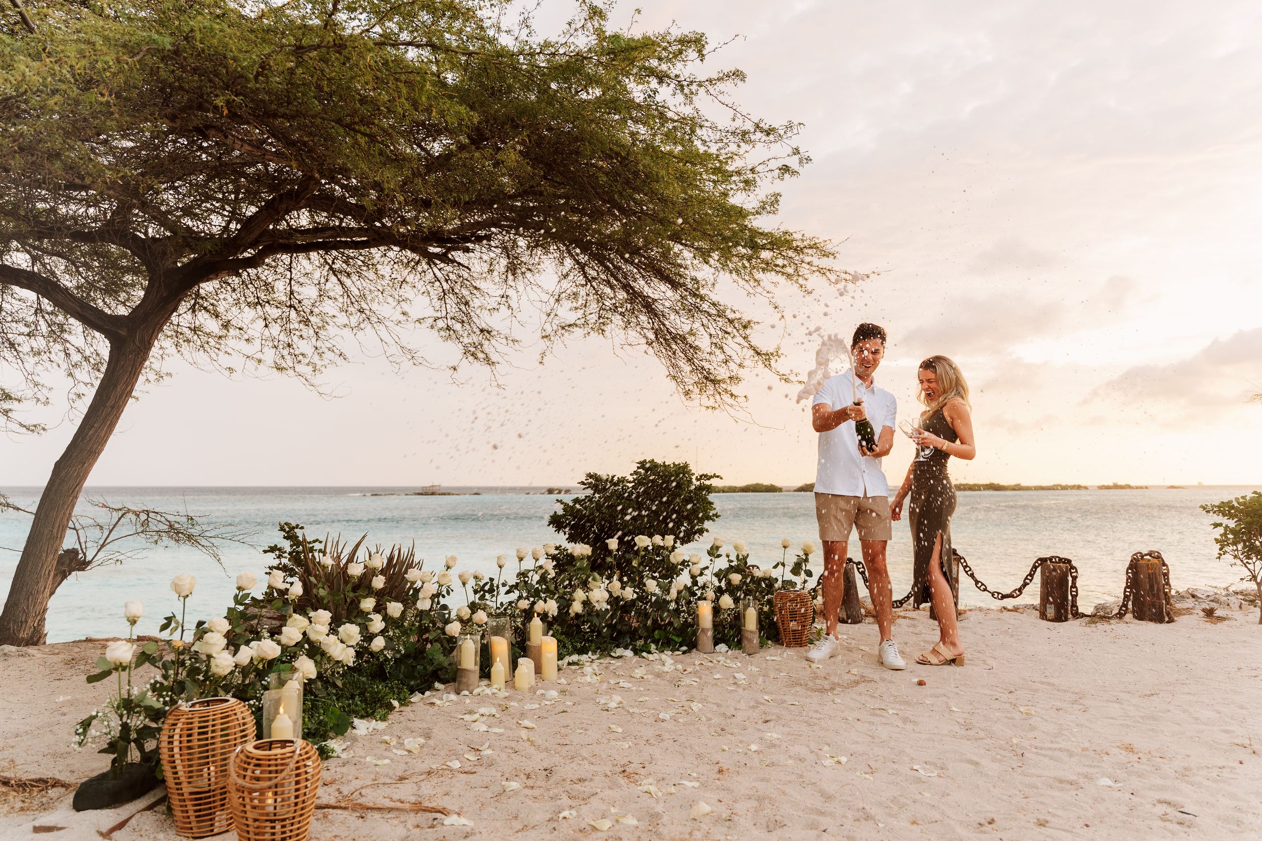 Couple celebrating a romantic beach proposal at sunset with floral decorations, candles, and champagne on a tropical shore.