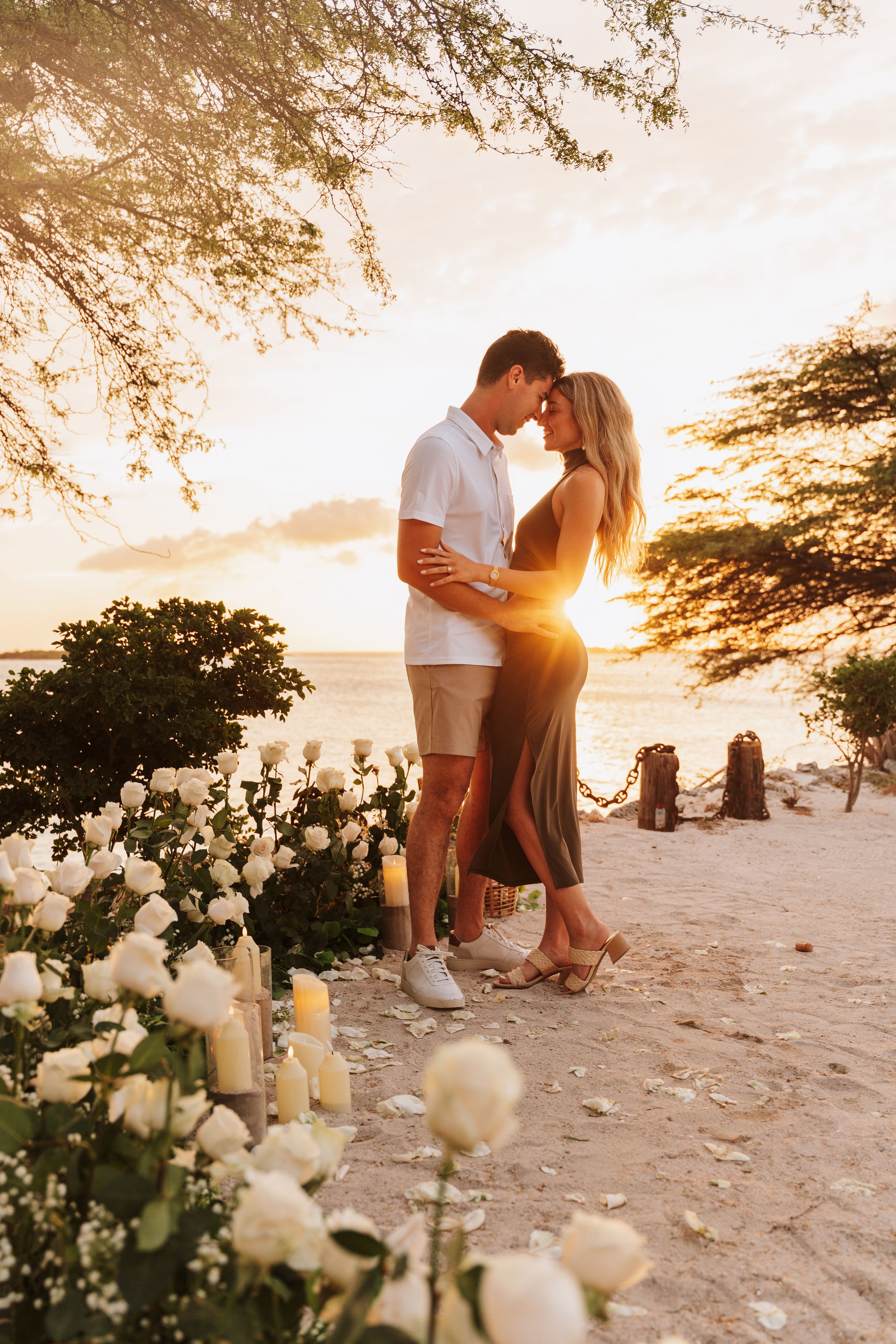 Romantic couple embracing at sunset on a sandy beach surrounded by white roses and candles.