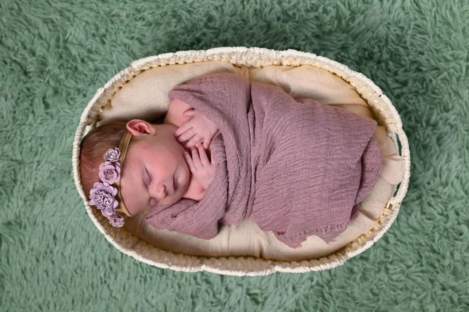 Newborn baby swaddled in pink wrap sleeping in a wicker basket with a floral headband on green background.