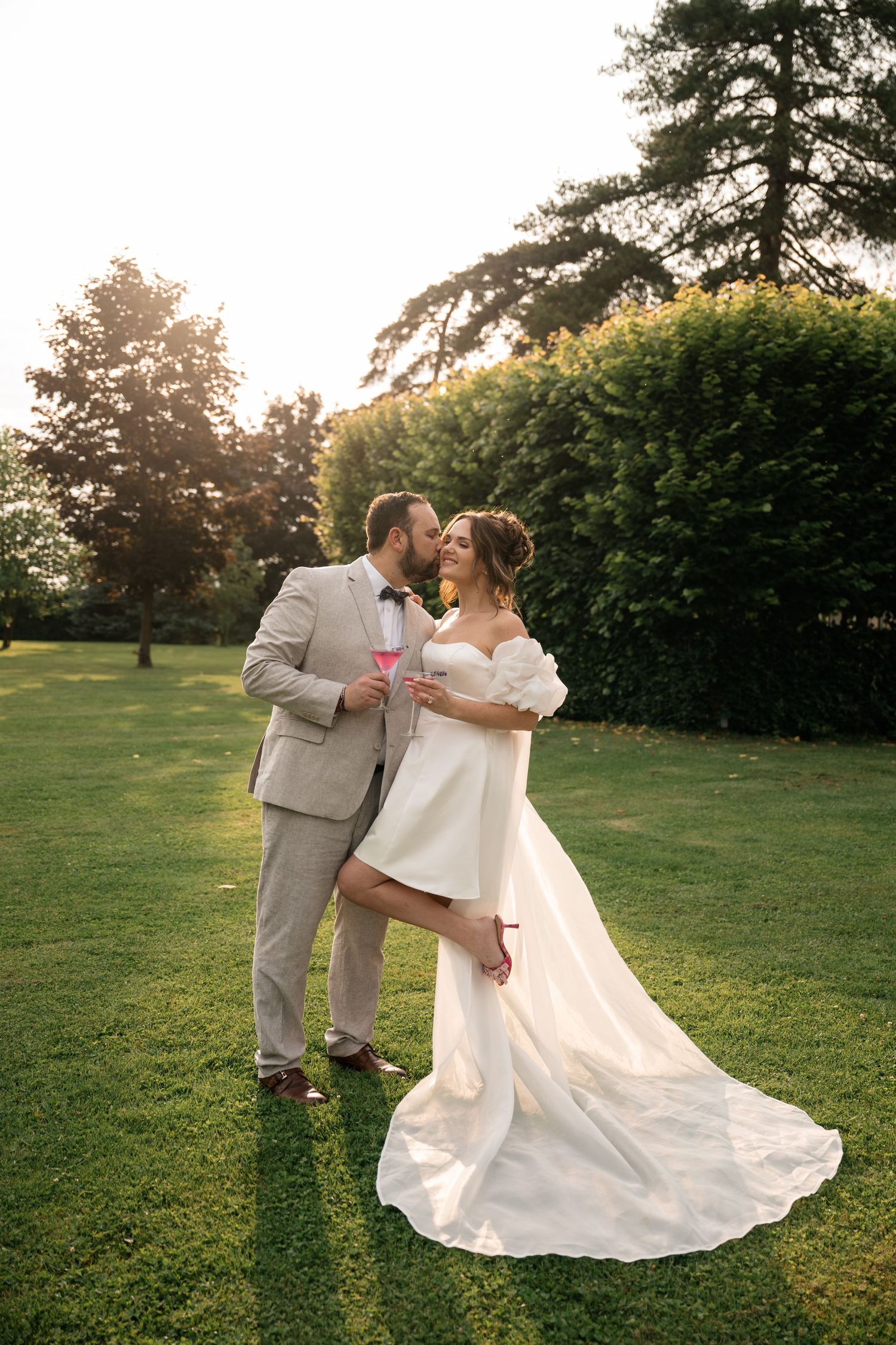 A romantic wedding couple shares a tender moment at sunset on a lush green lawn surrounded by tall trees.