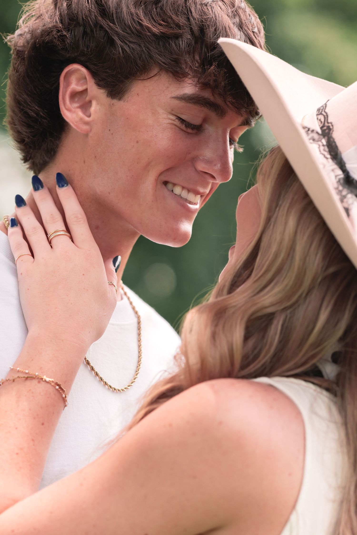 A smiling young couple sharing a romantic moment, woman wearing a cowboy hat and gold bracelet, man wearing a gold chain necklace.