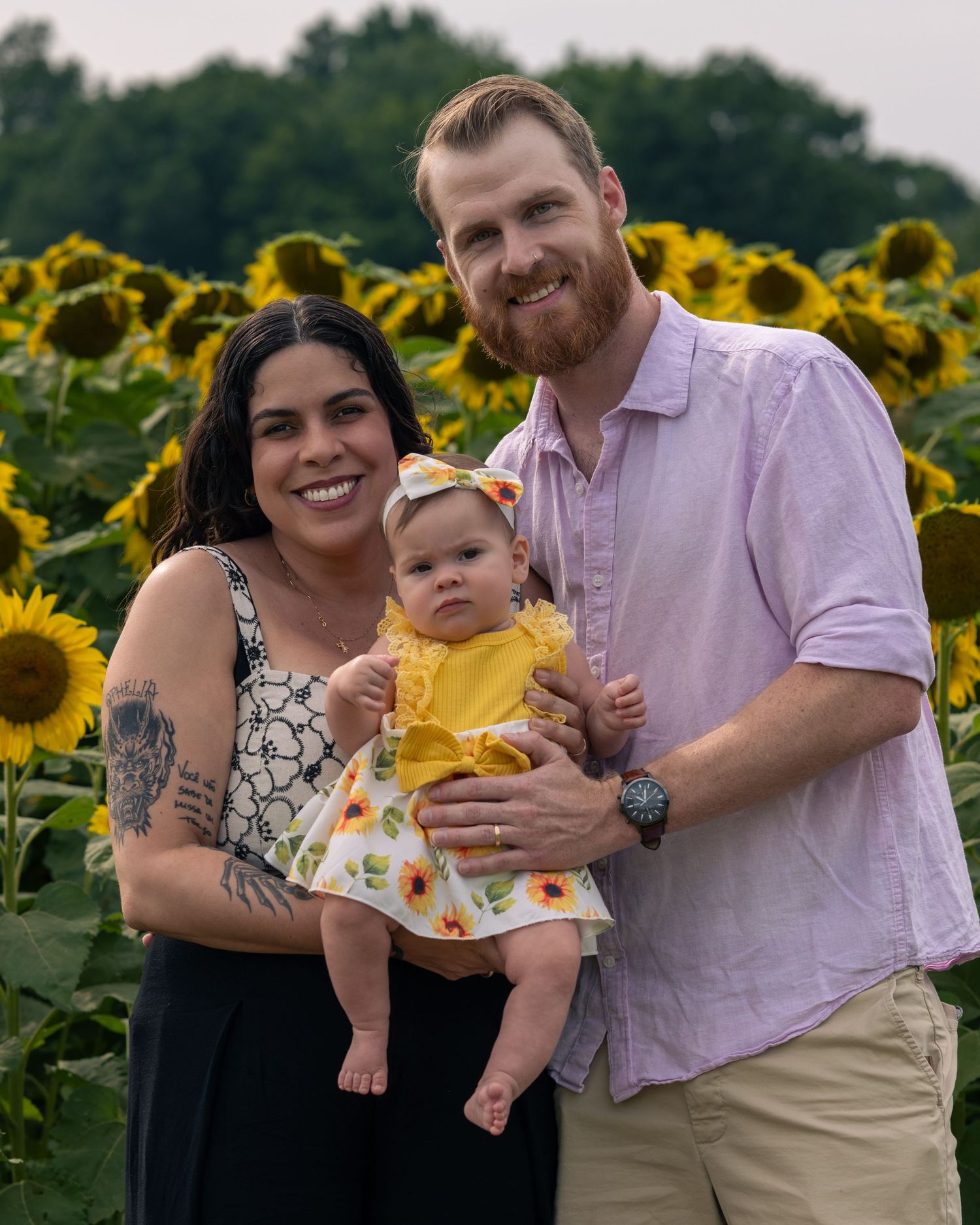 A smiling couple holds their baby girl in yellow dress amid a vibrant sunflower field backdrop.